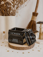 Decorative black box with text on a wooden surface with dried flowers and a broom in the background.